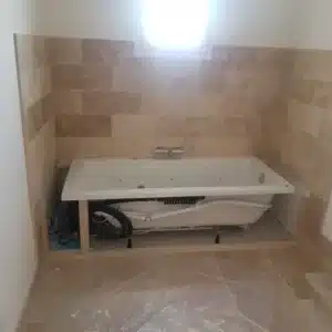 Installation d'une baignoire balnéo encastrée sur sol carrelé, murs revêtus de carreaux de travertin clairs.