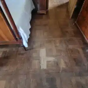 Parquet ancien en bois foncé usé avec une marque de croix claire au centre, près d'un lit à baldaquin.