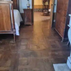 Parquet ancien à motif en losange dans une chambre meublée de meubles en bois foncé, menant à un couloir.