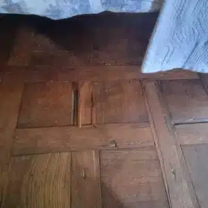 Plancher ancien en parquet de chêne verni marron sous un lit, montrant des joints usés et de légers dommages.